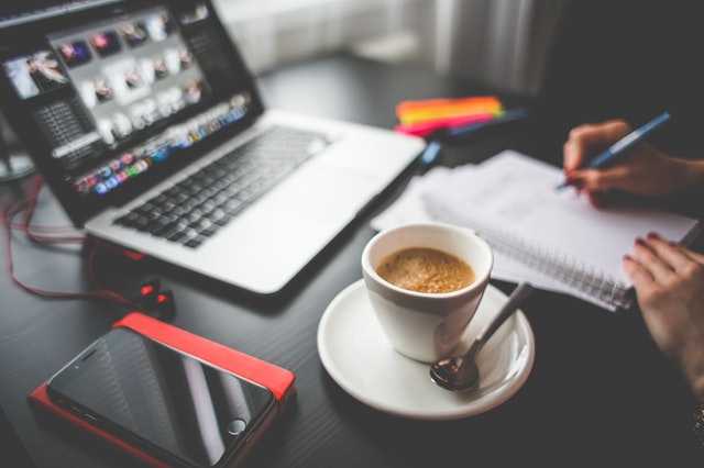 Desk with computer, coffee, person writing in notebook
