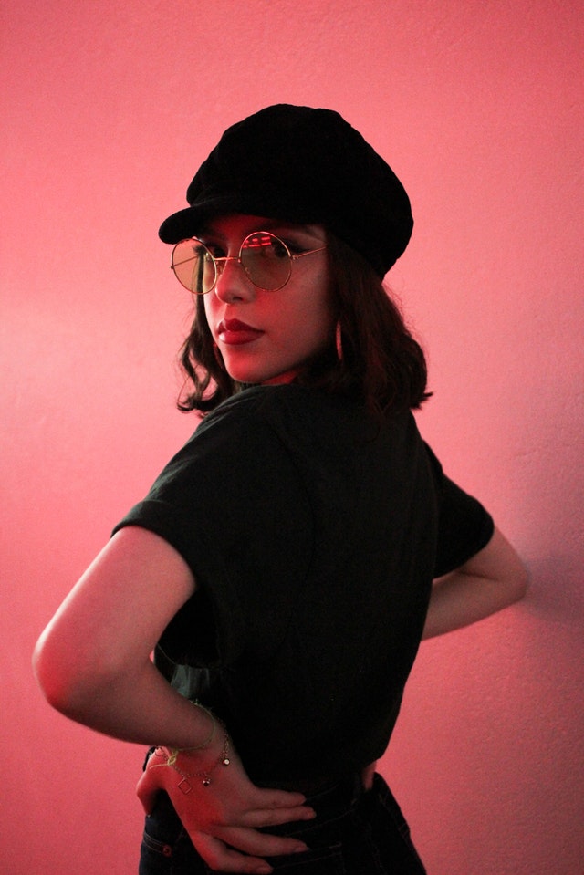 Woman in retro all black outfit with mod hat and glasses in front of pink wall
