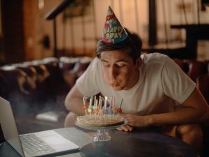 Man in party hat blowing out candles on birthday cake in front of laptop webcam