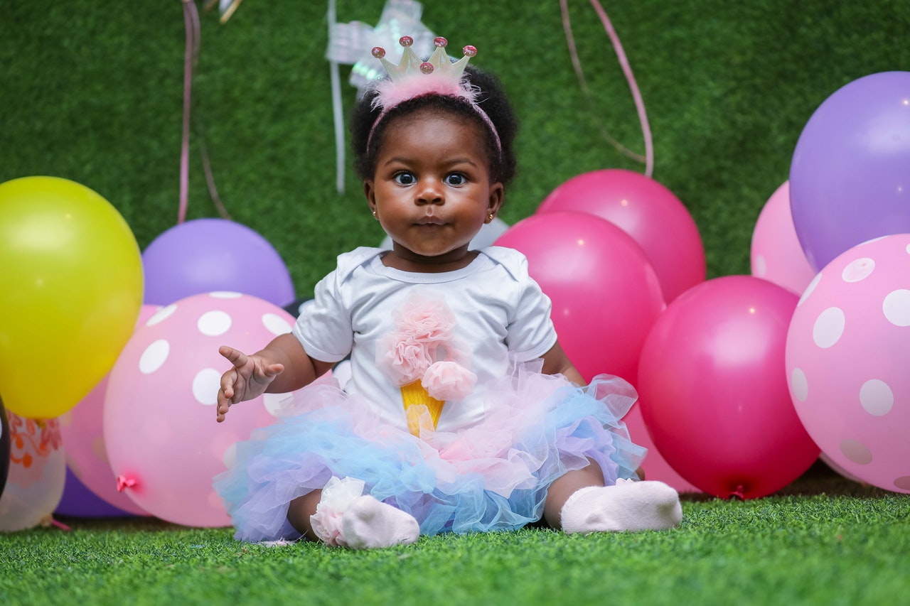 Adorable baby in birthday dress and crown sitting in front of balloons