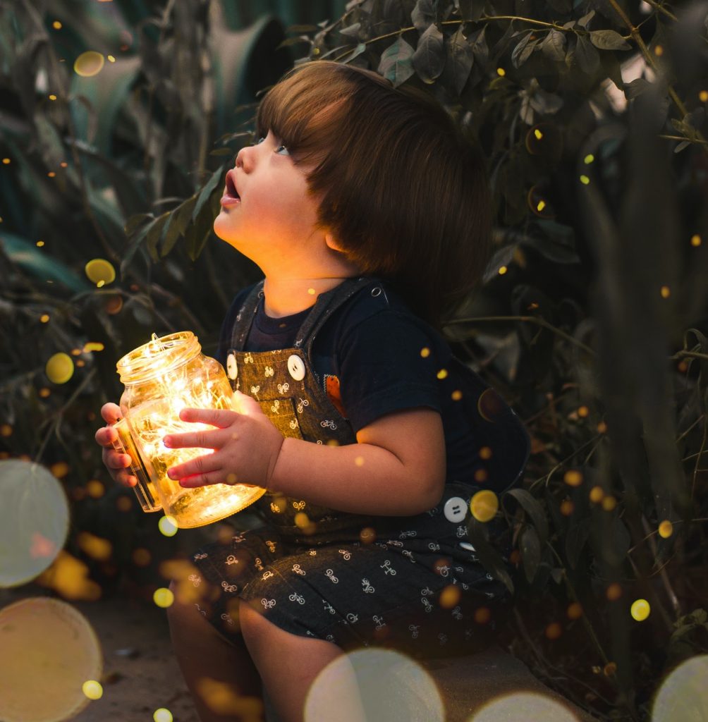 Cute kid in summer overalls holding jar of light in front of plants